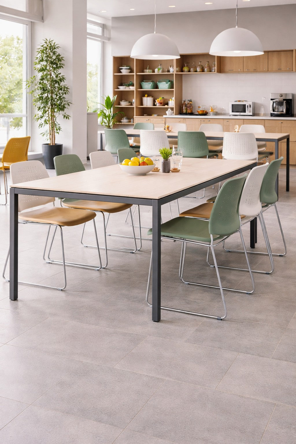 Une cuisine moderne et lumineuse présente une longue table de collectivité Orba - Plateau mélaminé de Monsieur Bureau, entourée de chaises vertes et beiges. Des étagères ouvertes, des appareils électroménagers, des plantes et de grandes fenêtres apportent beaucoup de lumière naturelle.