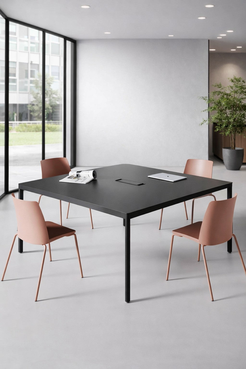 Dans une salle de réunion moderne, Monsieur Bureau Table de réunion Black Design 120x120 cm & Top Access, quatre chaises roses, un ordinateur portable et des documents. De grandes fenêtres inondent la pièce de lumière et une plante en pot trône dans un coin.