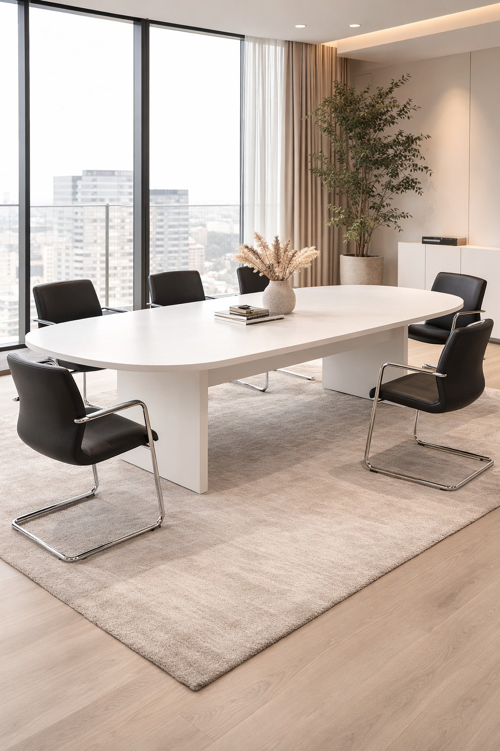 Une salle de conseil moderne avec une Table de réunion ovale Olympe 12 à 24 personnes de Monsieur Bureau, six chaises noires, un tapis beige, de grandes fenêtres donnant sur la ville, une plante en pot et un vase avec des fleurs séchées sur la table.#finitions_blanc