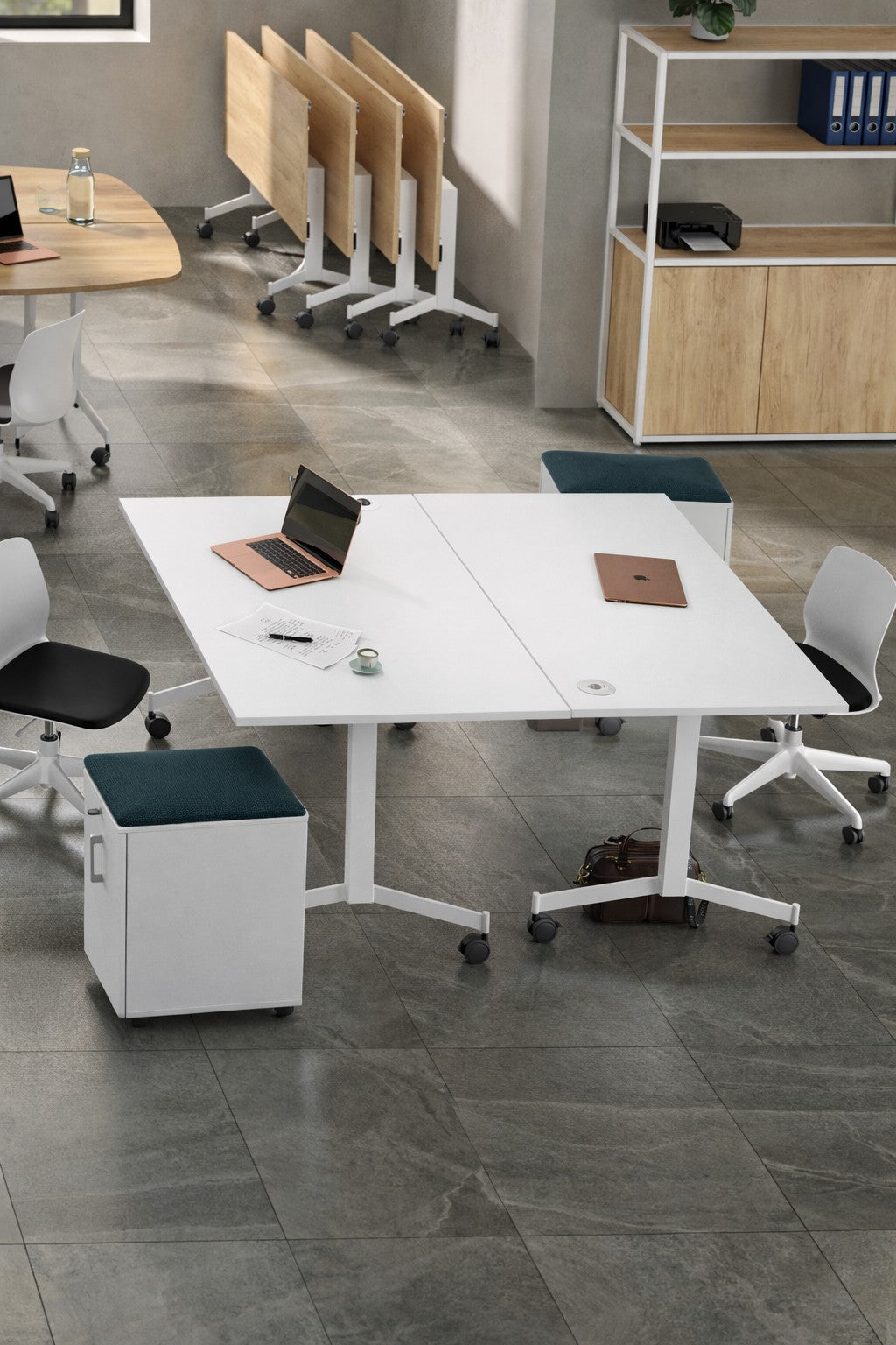 Un bureau moderne avec des tables blanches, des chaises à roulettes, des ordinateurs portables, des cahiers, une tasse et la Table Réunion Abattante et Mobile Quick Moove de Monsieur Bureau sur un sol carrelé gris. Des étagères et des bureaux supplémentaires apparaissent à l'arrière-plan.#combo_blanc_blanc