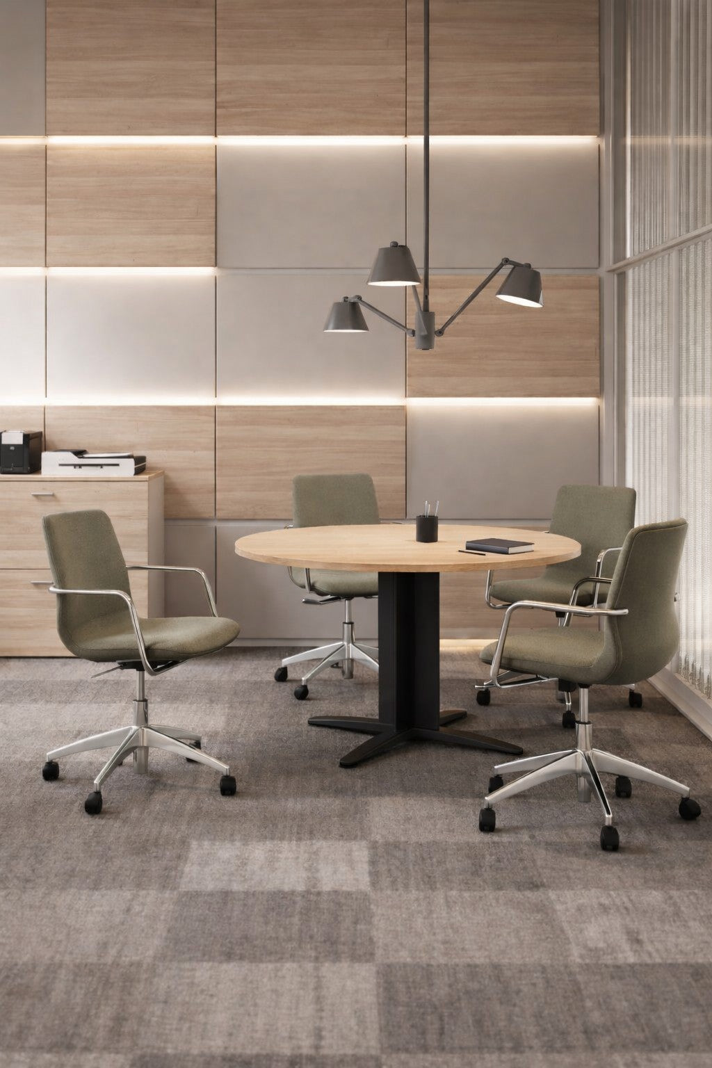 Un espace collaboratif moderne comprenant un Monsieur Bureau Table de réunion ronde Atlas (diamètre 116 cm), quatre chaises pivotantes vertes, une lampe suspendue au-dessus, des murs lambrissés et une armoire de fond avec des fournitures de bureau.#finitions_noir_chene_moyen