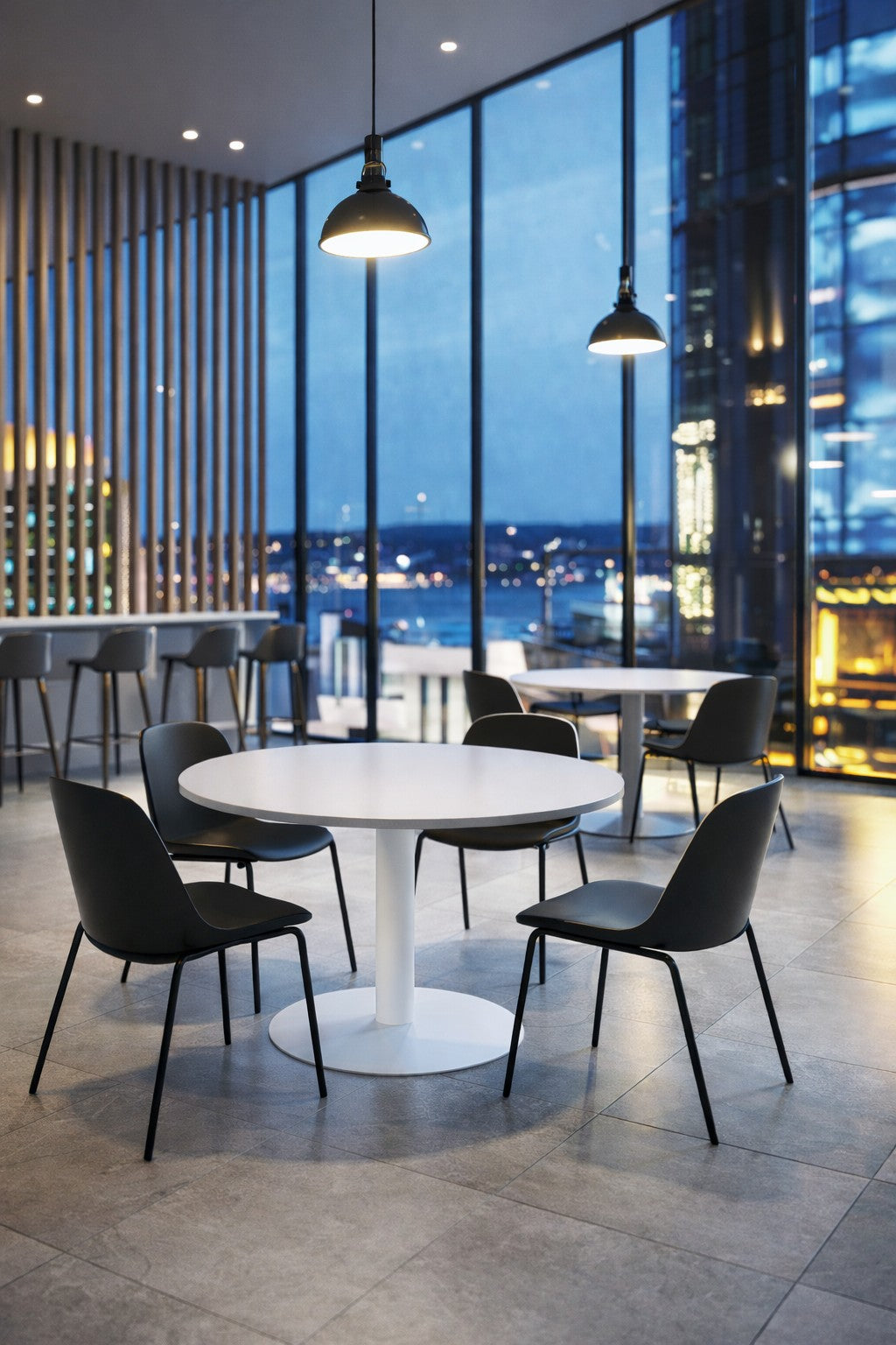 Salon moderne avec la Table Réunion Ronde Sevka (Ø100/Ø120) de Monsieur Bureau et des chaises noires près de fenêtres allant du sol au plafond et donnant sur la ville au crépuscule. Des lampes suspendues réchauffent l'espace confortable et contemporain.#combo_blanc_blanc