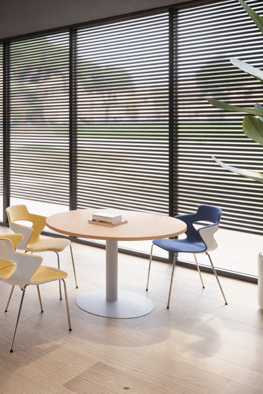 Un espace de réunion moderne avec un Monsieur Bureau Table Réunion Ronde Sevka 4 Pers (Ø100/Ø120), trois chaises jaunes, une chaise bleue, des livres sur la table, de grandes fenêtres avec des stores horizontaux, du mobilier professionnel, et de la lumière naturelle.#combo_chene_fil_blanc