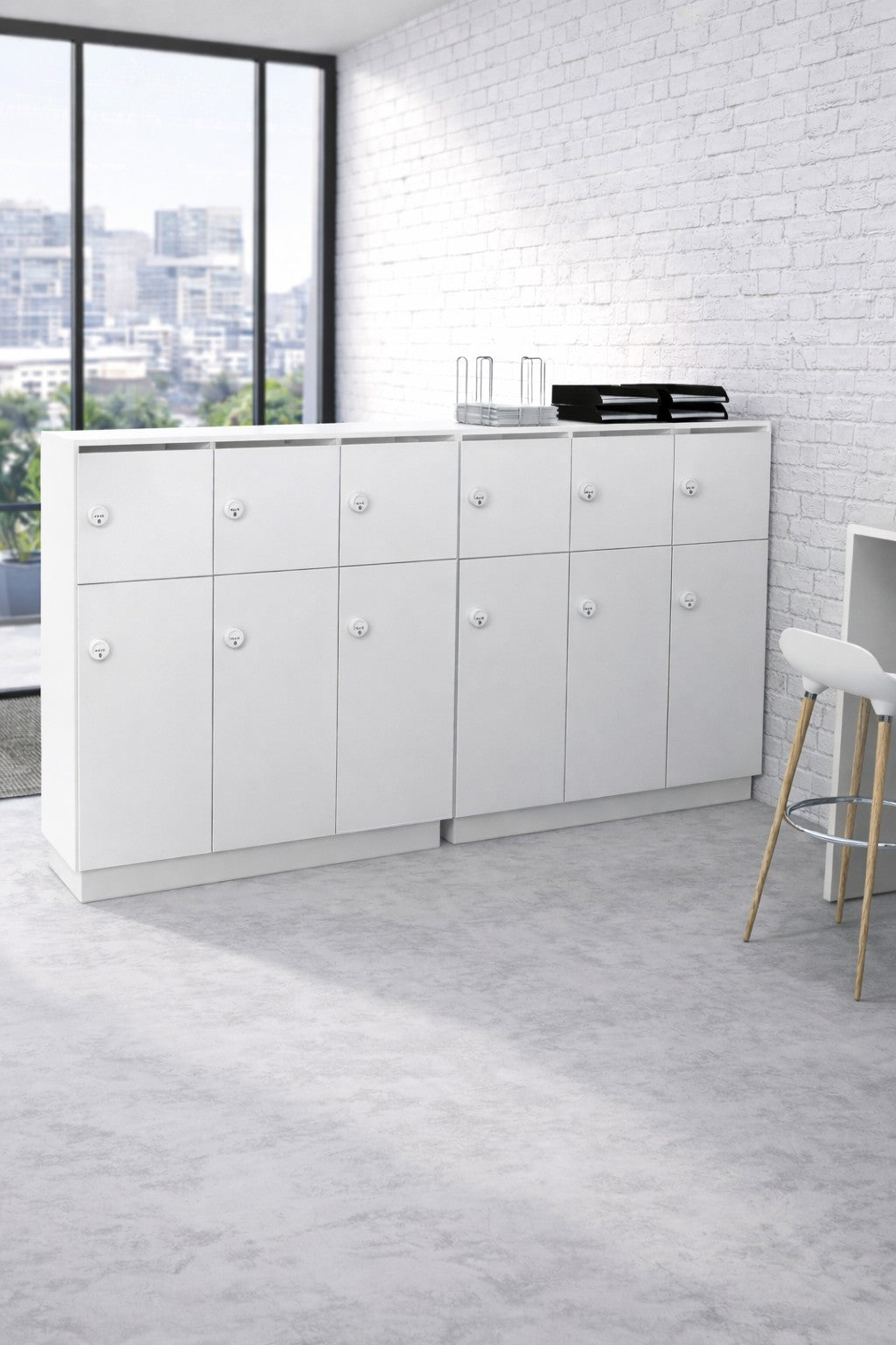 Un bureau moderne avec deux armoires blanches et un Monsieur Bureau Vestiaire Bois 3 Colonnes Astral sur Socle contre un mur de briques blanches, contenant des verres, des plateaux et des dossiers. Un tabouret haut est posé à proximité, avec de grandes fenêtres encadrant le paysage urbain.#combo_blanc_blanc