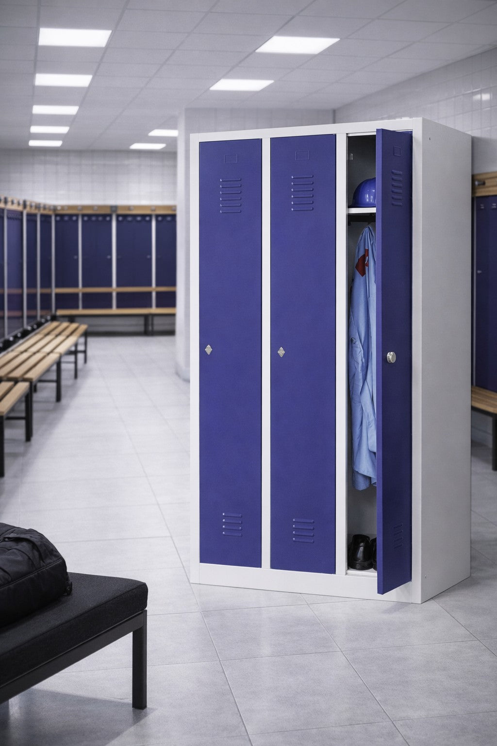 Un vestiaire spacieux comporte des casiers Vestiaire métallique Talos 1 porte de Monsieur Bureau (1-3 colonnes), des bancs et des sols carrelés. Un casier est ouvert, montrant une chemise bleue sur un cintre, un casque et des chaussures à l'intérieur ; la pièce est vide.