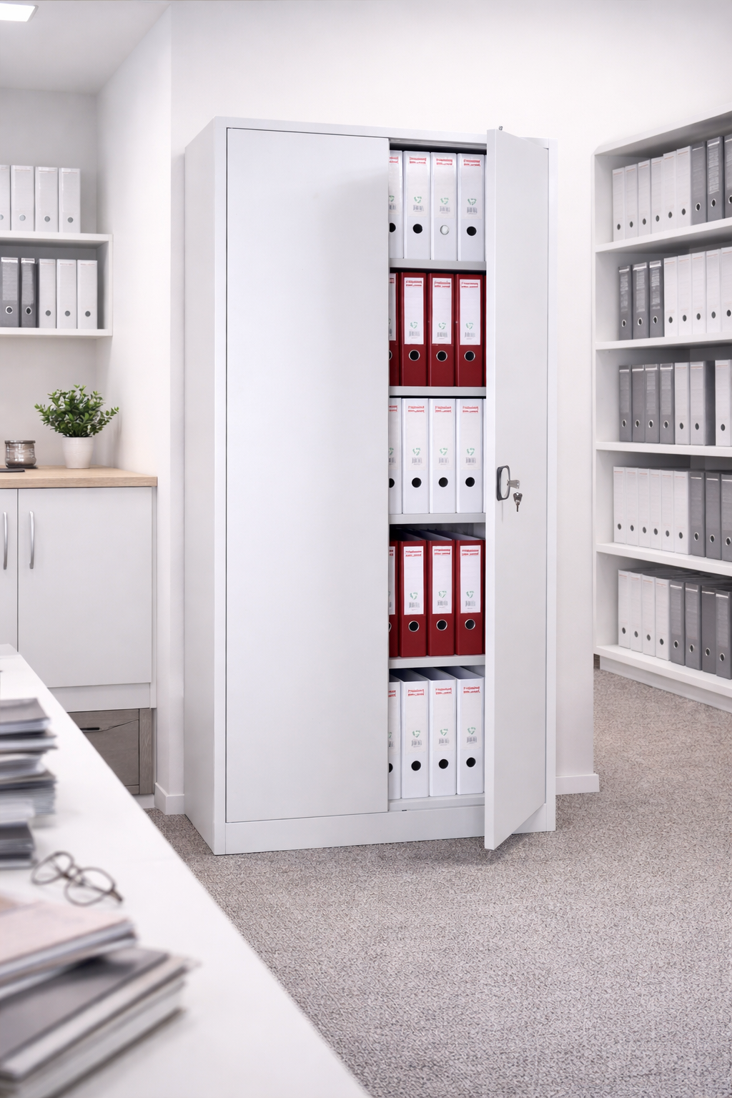 L'Armoire Haute Métal à portes battantes SOLID H180 cm de Monsieur Bureau est ouverte et présente sur ses étagères des classeurs blancs et rouges bien rangés. D'autres classeurs et une petite plante en pot décorent le comptoir voisin.