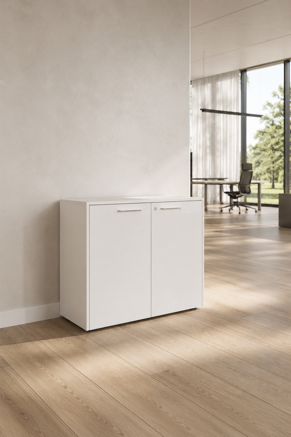 L'Armoire basse Passe Partout 2 portes de Monsieur Bureau (L90 x H79.5) se trouve contre un mur clair dans un bureau spacieux avec de grandes fenêtres, des planchers en bois et un bureau vide à l'arrière-plan.#finitions_blanc