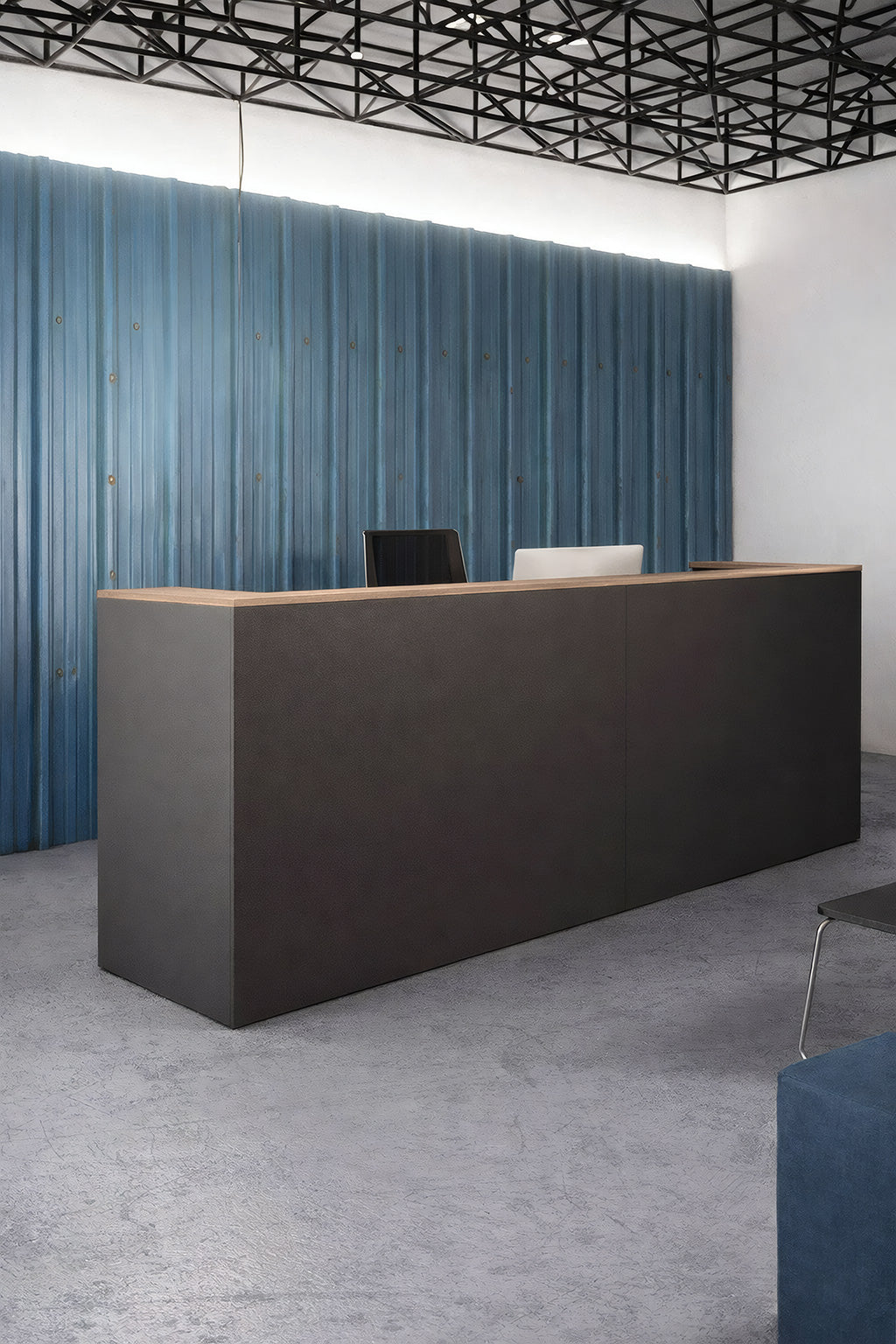 L'espace d'accueil d'un bureau moderne comprend un Monsieur Bureau Comptoir d'accueil professionnel Delta Pro L145 à L365 dans un style minimaliste sombre, deux chaises derrière lui, un mur texturé bleu, un sol en béton et un plafond à grille géométrique noire.#finitions_Graphite_avec_top_Chêne_moyen_199