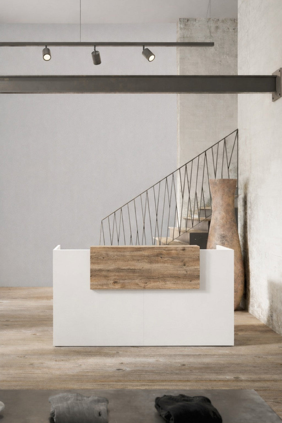 Un intérieur minimaliste présentant la Banque d'accueil professionnelle droite ABBA de Monsieur Bureau en blanc et en bois, posée sur un sol en béton avec un vase rustique, un escalier à rampe métallique et un éclairage sur rail épuré sur un mur texturé gris clair.#combo_timber_blanc