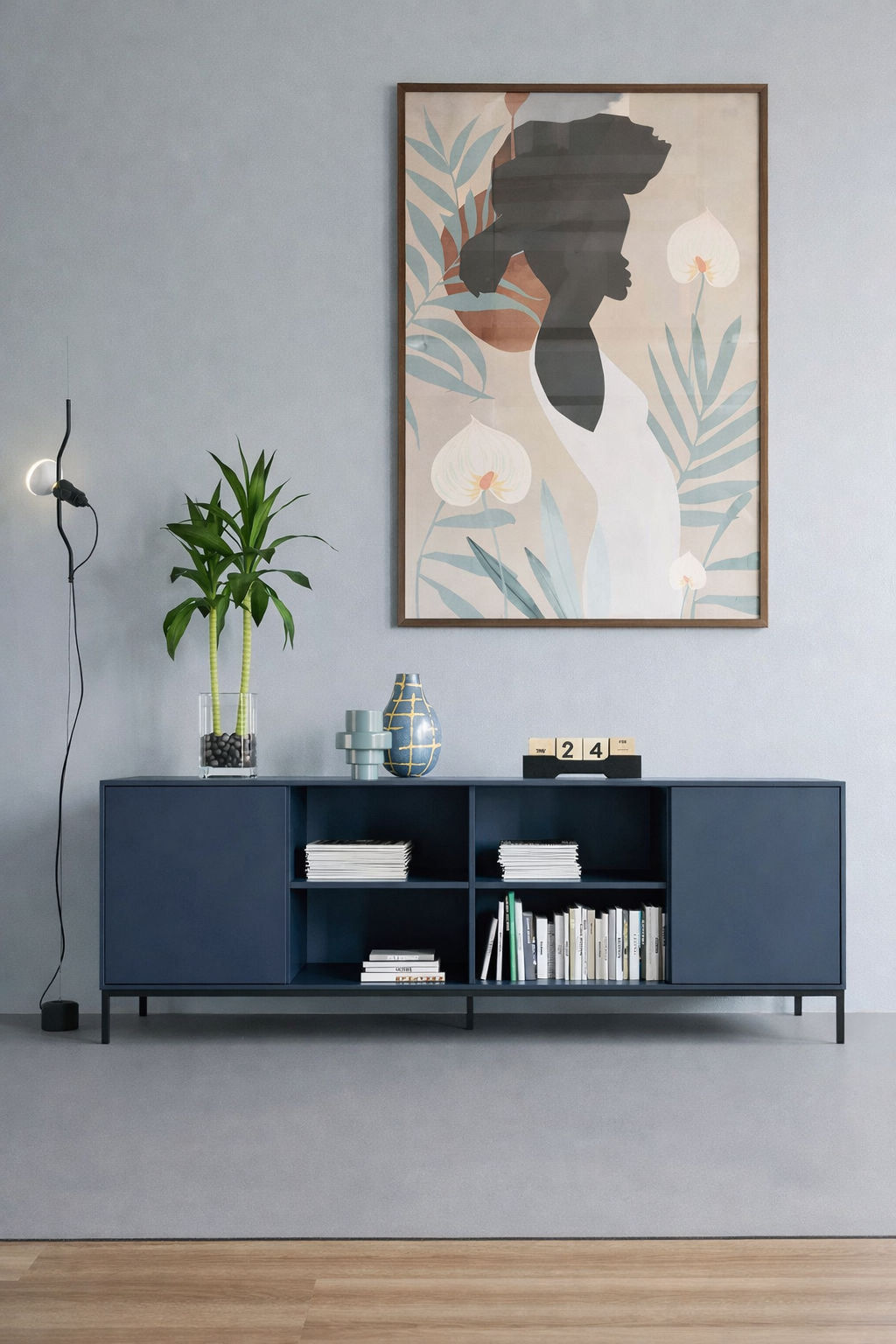 Le Buffet Quadra - 2 portes et 4 niches de Monsieur Bureau en bleu présente des livres, de la décoration et une plante. Un tableau abstrait représentant une silhouette de femme est suspendu au-dessus, tandis qu'une applique murale minimaliste sur la gauche rehausse l'ambiance moderne d'inspiration italienne.