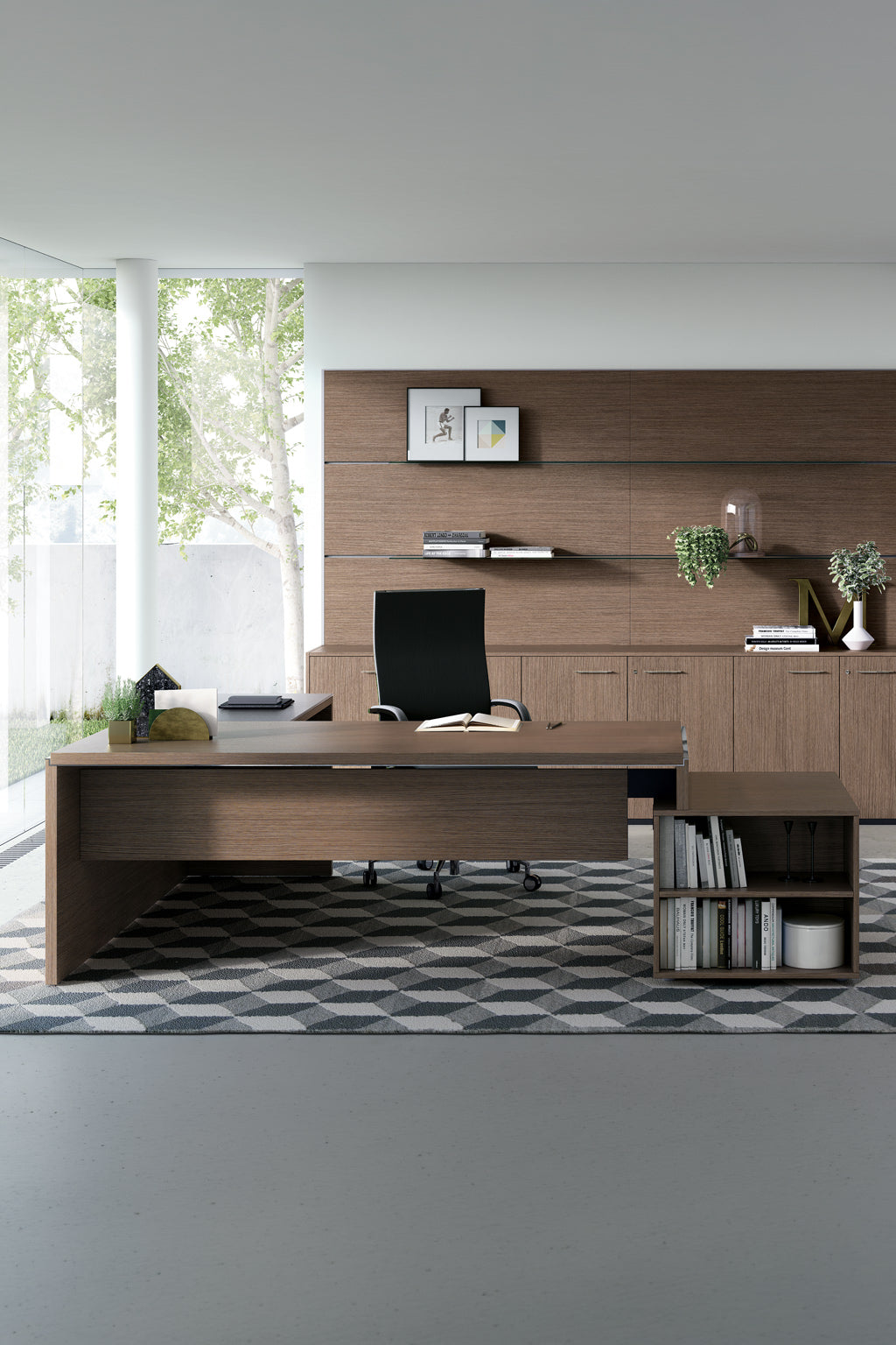 Bureau moderne comprenant le Monsieur Bureau "Bureau de direction Asset avec retour et rangement", une chaise noire, des livres, des plantes, un tapis géométrique et des étagères murales avec de la décoration. De grandes fenêtres laissent entrevoir les arbres à l'extérieur, remplissant l'espace de lumière naturelle.