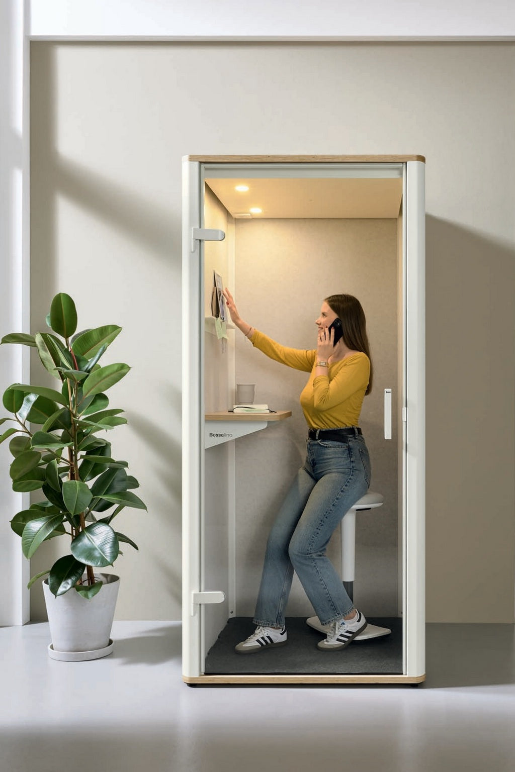 Une femme vêtue d'un haut jaune et d'un jean parle au téléphone à l'intérieur de la Cabine acoustique Silen - Phone booth 1 personne de Monsieur Bureau, en attrapant un livre. Une plante en pot à l'extérieur agrémente cet espace de concentration confortable.
