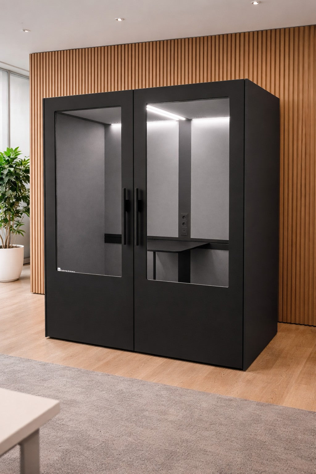 La cabine Monsieur Bureau Cabine Acoustique Double No Sound Standard 2 pers, de couleur noire moderne avec des portes en verre, est posée sur un plancher en bois à côté d'un mur lambrissé. Elle dispose d'un éclairage intérieur et d'un bureau intégré pour deux personnes.#finitions_noir