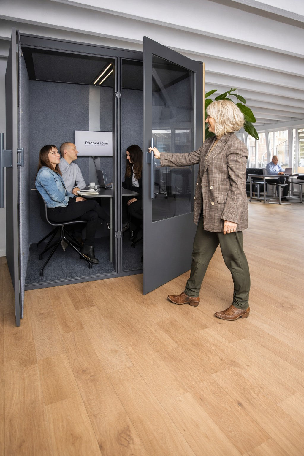 Une femme ouvre la porte d'un Monsieur Bureau Cabine Acoustique Réunion No Sound Standard 4 pers, où trois personnes se réunissent à l'intérieur de cette cabine de bureau moderne avec des planchers en bois et de grandes fenêtres.