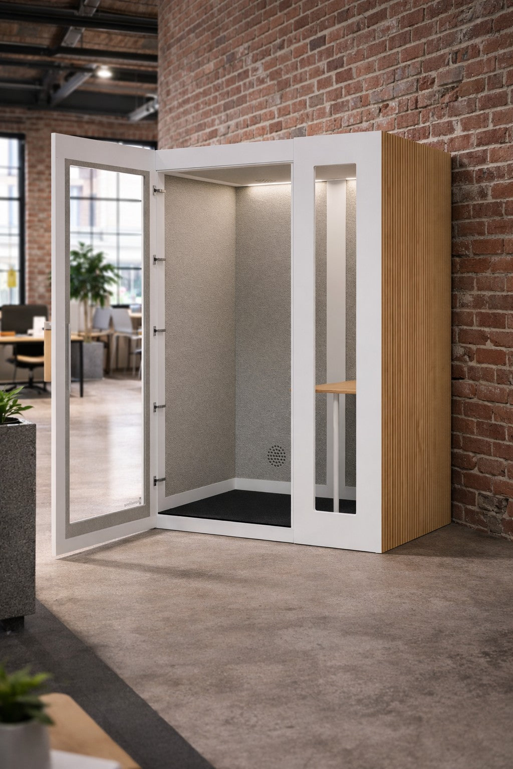 La Cabine acoustique de Travail NO SOUND Executive 1 pers de Monsieur Bureau est dotée d'une porte en verre, d'un intérieur gris clair, d'une petite étagère et de lattes latérales en bois, offrant une isolation acoustique dans les bureaux ouverts avec des murs en briques apparentes.#finitions_blanc