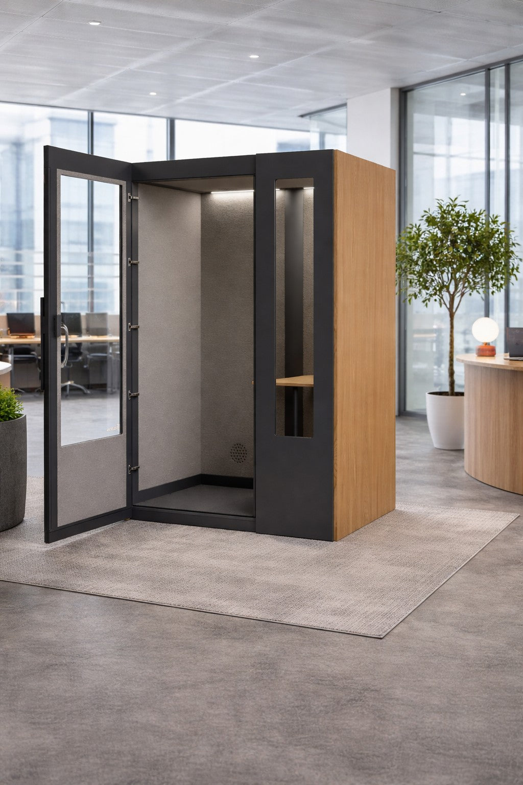 La Cabine acoustique de Travail NO SOUND Standard 1 pers de Monsieur Bureau se compose de panneaux en bois et en noir avec une porte en verre, posés sur une moquette au milieu de meubles de bureau et de plantes - idéale comme cabine téléphonique pour les bureaux en open space.#finitions_chene
