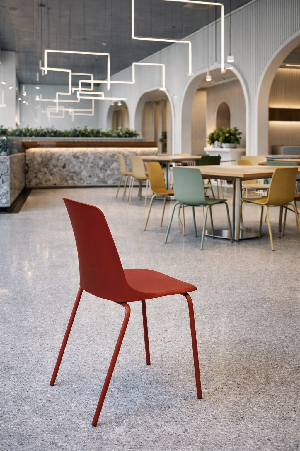 Un intérieur moderne mettant en valeur la Chaise empilable ALIX de Monsieur Bureau au premier plan, avec des chaises et des tables pastel derrière, des détails muraux arqués, des plafonniers géométriques et un style mobilier professionnel minimaliste.