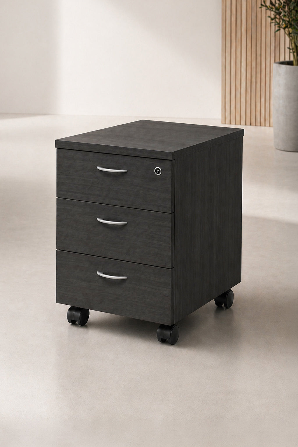 Un Caisson mobile Budget / Effect 3 tiroirs de Monsieur Bureau, doté de poignées en argent, est posé sur un sol clair dans une pièce moderne et minimale, avec des rangements intelligents et une plante en pot en arrière-plan.#combo_frene_noir_3_tiroirs