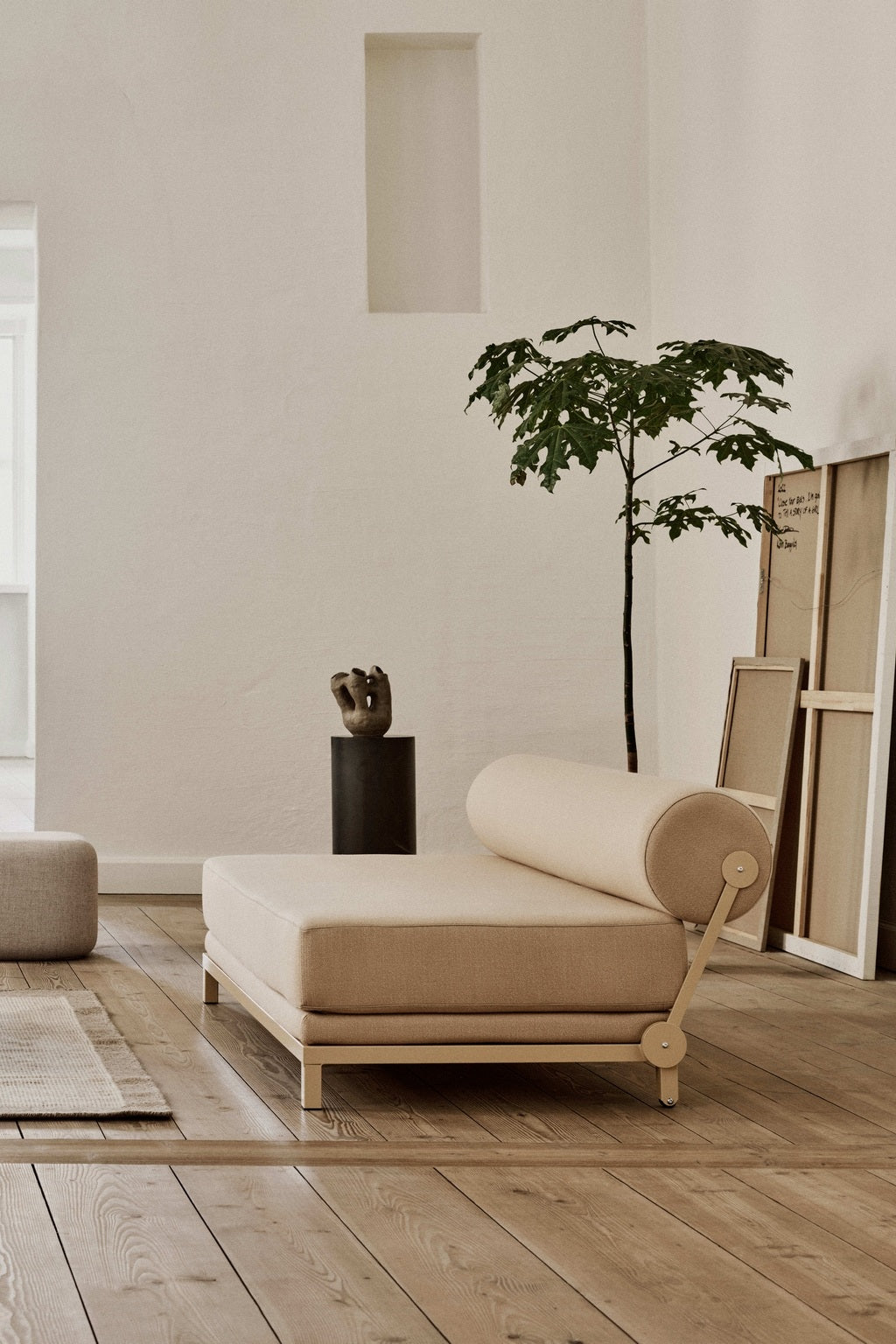 Un salon minimaliste présente le Canapé-Lit Nyx de BUSK+HERTZOG pour Monsieur Bureau, de couleur beige, posé sur un sol en bois clair, avec une plante en pot et un tableau encadré contre un mur blanc, baigné de lumière naturelle.