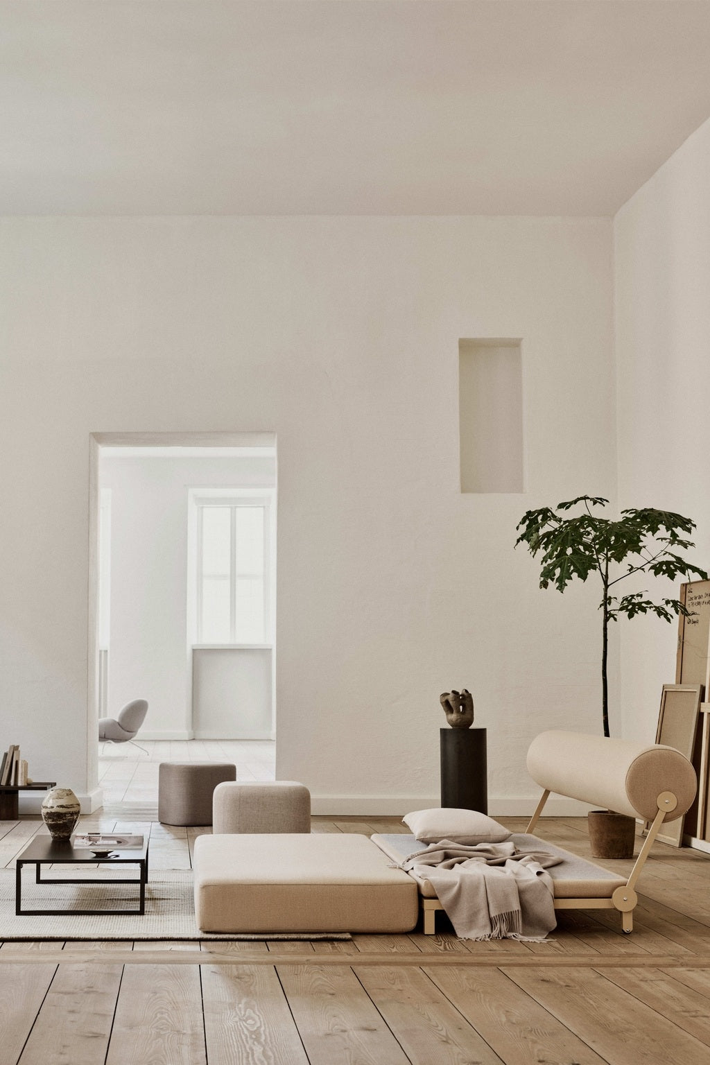 Salon minimaliste avec des planchers en bois clair, des murs blancs et de grandes fenêtres. Le Canapé-Lit Nyx de BUSK+HERTZOG pour Monsieur Bureau ajoute un style moderne à un décor neutre, une plante verte et des objets décoratifs pour une atmosphère aérée.