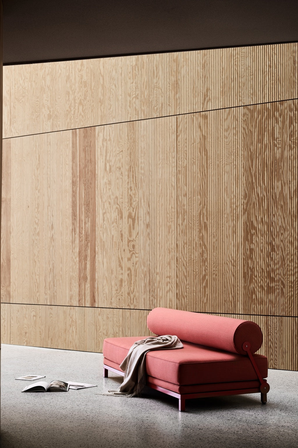 Un intérieur moderne avec le Canapé-Lit Nyx de BUSK+HERTZOG pour Monsieur Bureau, présenté avec des murs en bois clair, une chaise longue rose avec un oreiller cylindrique, une couverture drapée beige et un magazine ouvert sur le sol moucheté.#finitions_rouge