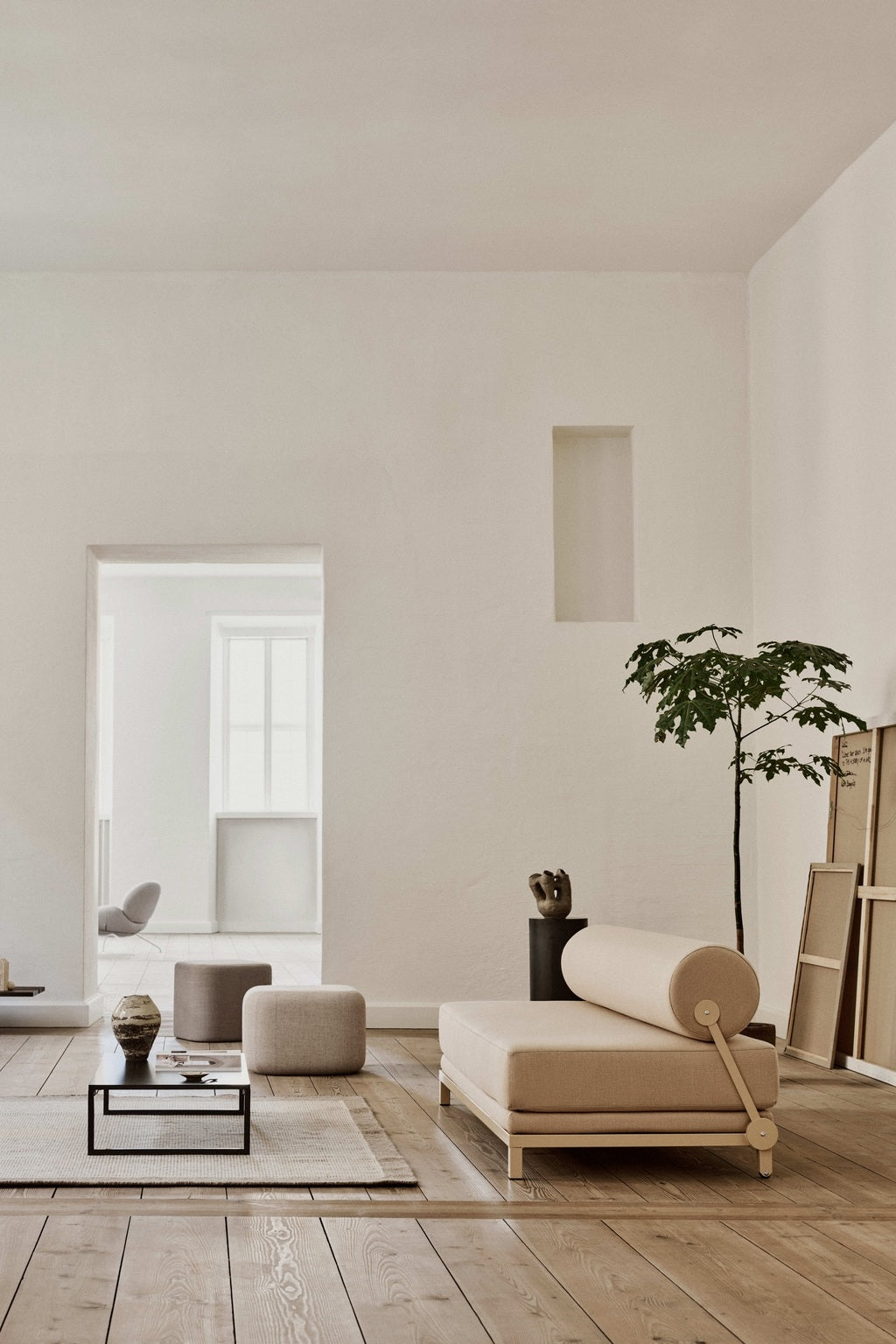 Salon minimaliste avec des planchers en bois clair, le Canapé-Lit Nyx de BUSK+HERTZOG (Monsieur Bureau), un décor moderne neutre, un petit arbre dans un pot noir, des œuvres d'art encadrées contre le mur, et de grandes fenêtres qui remplissent l'espace de lumière naturelle.#finitions_beige