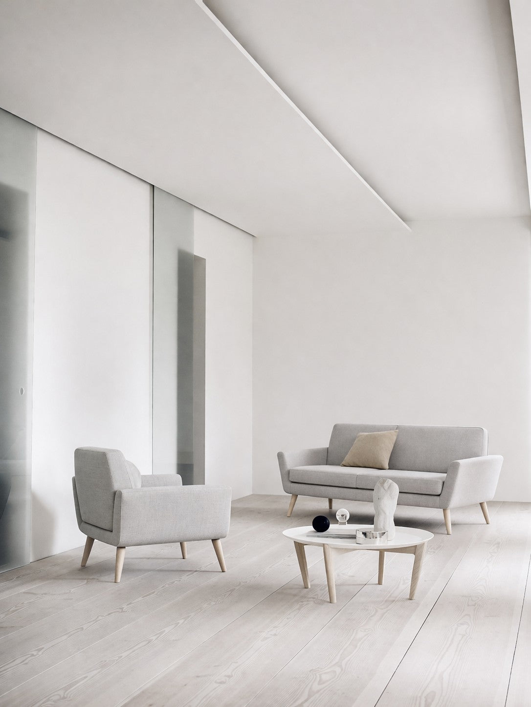 Salon minimaliste avec des planchers en bois clair, un Canapé scandinave Natt 2 places de Robert ZOLLER de Monsieur Bureau, un fauteuil et une petite table basse ronde blanche avec une sculpture. De grands panneaux de verre et des couleurs neutres ajoutent un confort scandinave.