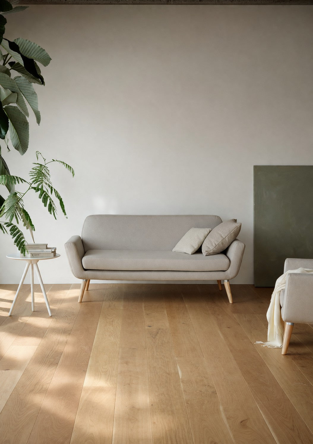 Un salon minimaliste avec le Canapé scandinave Natt de Robert ZOLLER (2 places) de Monsieur Bureau en gris clair, deux coussins beiges, une table d'appoint blanche avec un livre, une plante en pot verte et un sol en bois clair. La lumière naturelle met en valeur son confort scandinave.