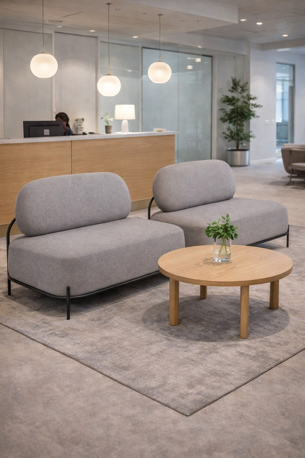 Un hall d'entrée de bureau moderne avec deux canapés Monsieur Bureau Brooklin 2 places, une table basse ronde en bois avec une petite plante dans un vase, et un bureau d'accueil en arrière-plan, le tout éclairé par des lampes globe suspendues pour une ambiance vintage-moderne.