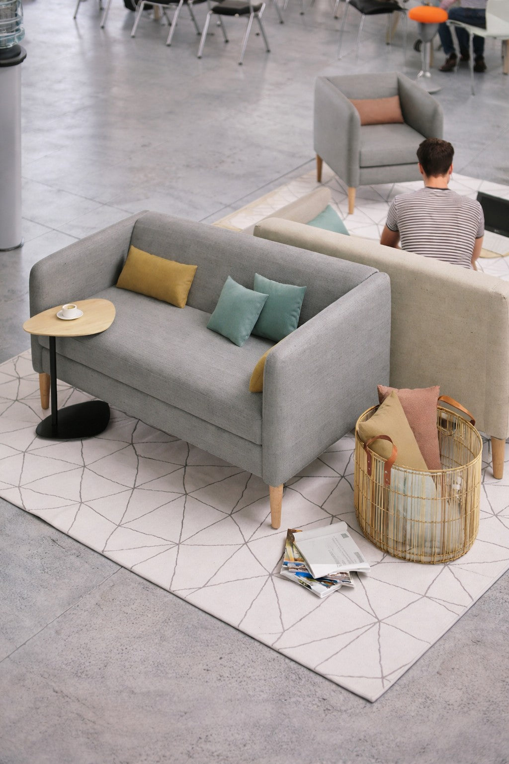 Un coin salon moderne présente le Canapé d'accueil Kanto pieds bois - 2 places de Monsieur Bureau en gris, des coussins colorés, une petite table d'appoint avec une tasse à café, un panier à revues et une personne assise sur le canapé, vue d'en haut.