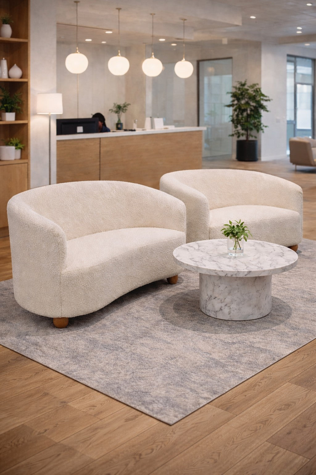 Deux canapés Canapé d'accueil Kurl 2 places tissu bouclette de Monsieur Bureau et une table basse ronde en marbre avec une plante sont posés sur un tapis gris dans un hall d'entrée de bureau élégant et bien éclairé avec des planchers en bois et un décor minimaliste.