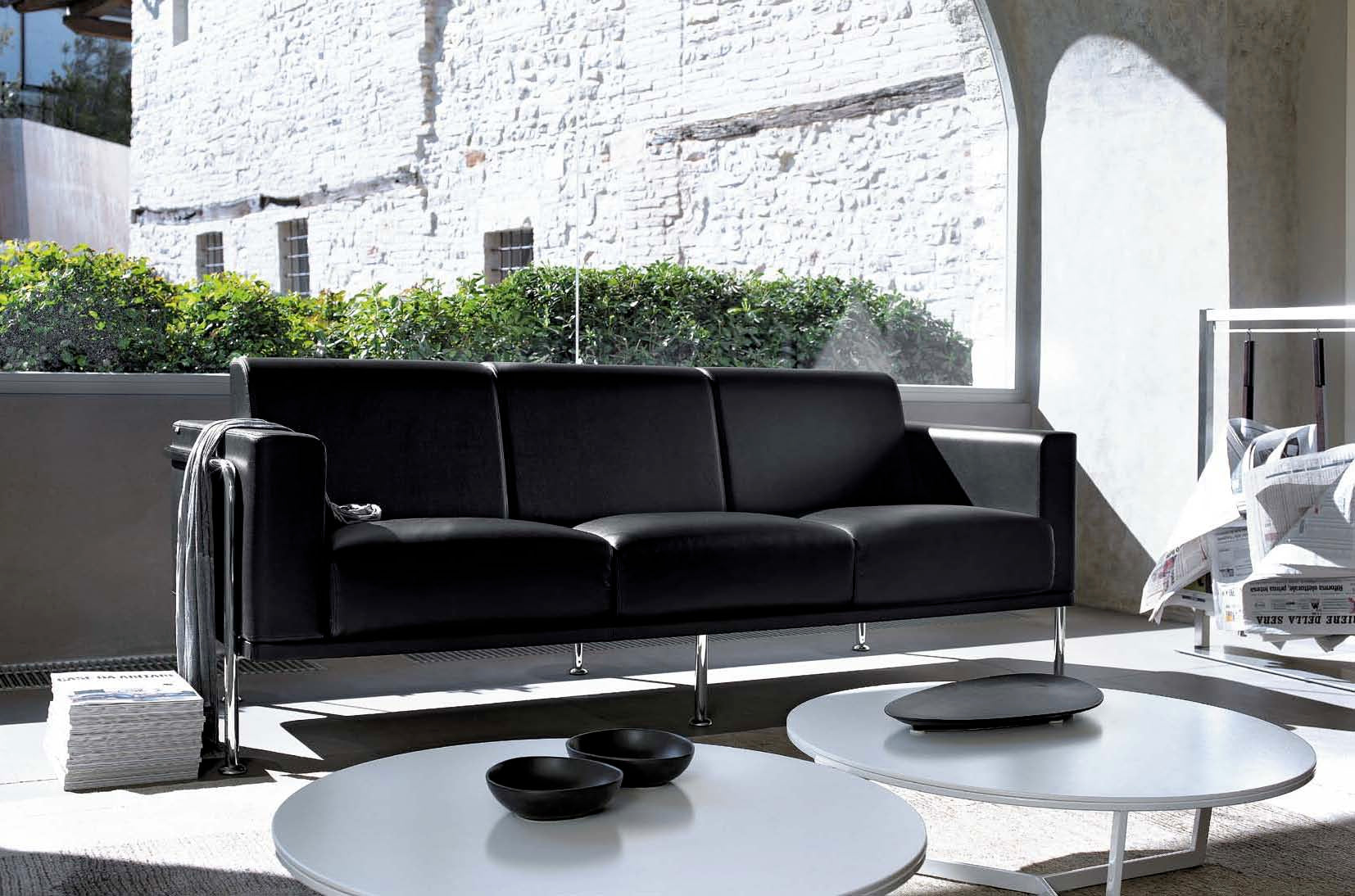 A Canapé d'accueil Matis en simili-cuir noir 2/3 places de Monsieur Bureau est installé dans une pièce lumineuse, près de deux tables basses rondes blanches décorées de bols noirs et d'une télécommande. Le soleil illumine un mur en pierre et la verdure extérieure.