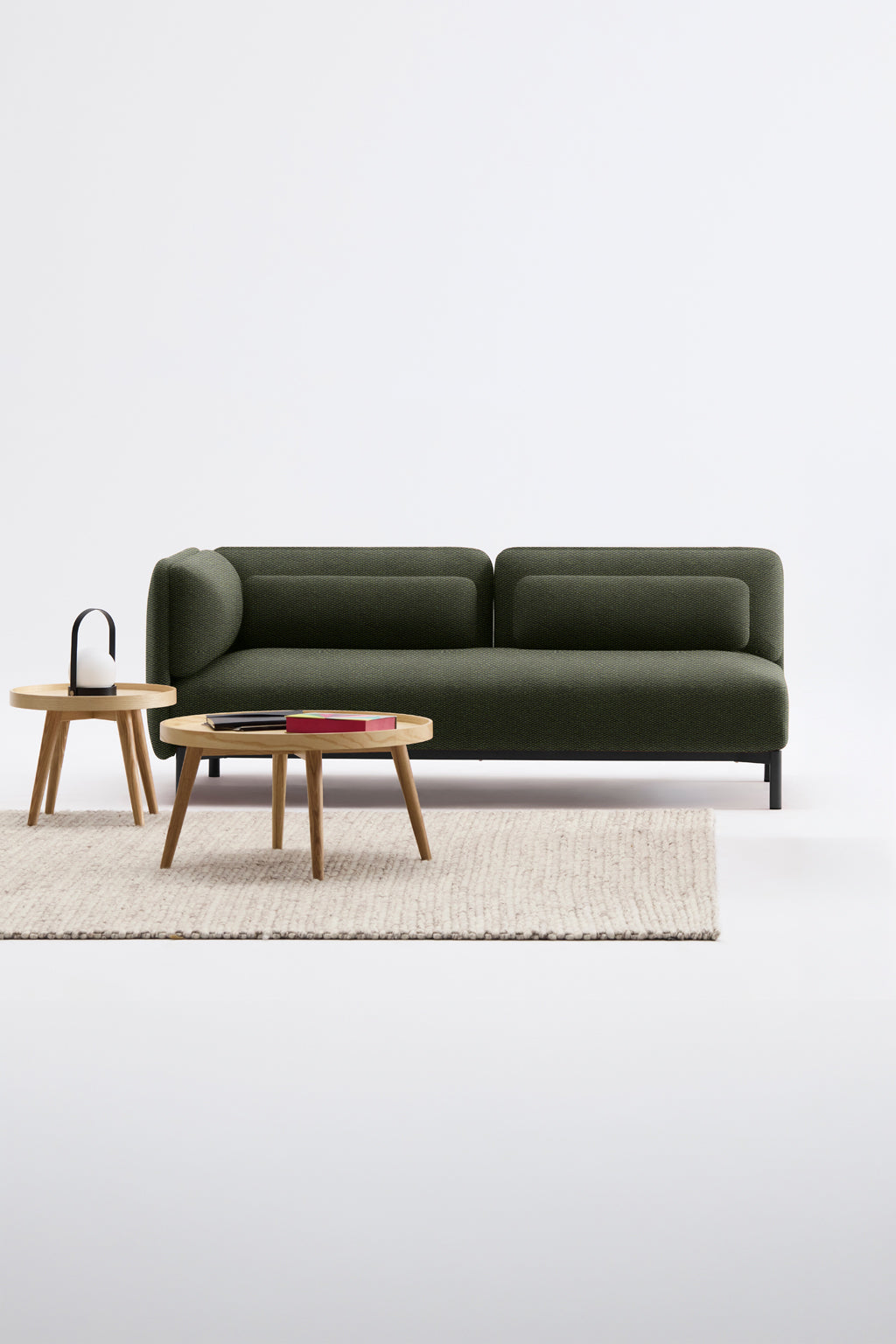 Le Canapé d'accueil modulaire Aurea - 1 accoudoir de Monsieur Bureau, d'un vert moderne, est posé sur un tapis à la texture claire entre deux tables rondes en bois - l'une avec des livres, l'autre avec un objet à poignée noire - sur un fond blanc uni.#finitions_vert