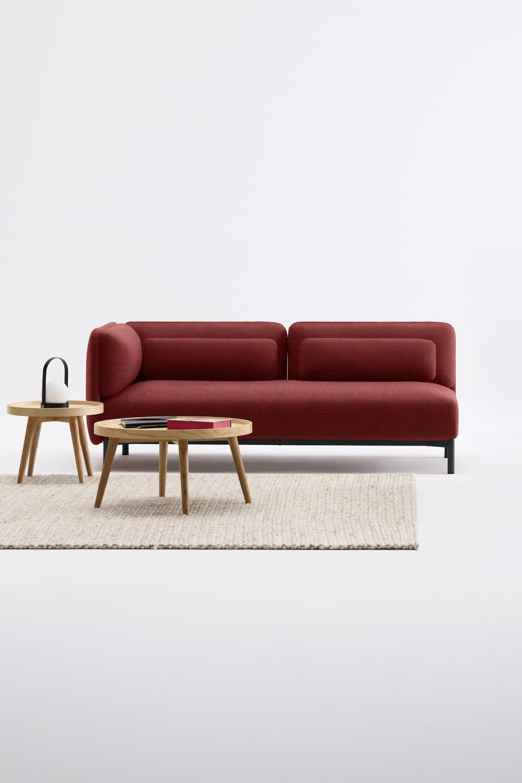 Le Canapé d'accueil modulaire Aurea - 1 accoudoir de Monsieur Bureau en rouge est posé sur un tapis à la texture pâle, avec deux tables basses en bois - l'une avec des livres, l'autre avec un ornement noir - sur un fond blanc.#finitions_terracotta