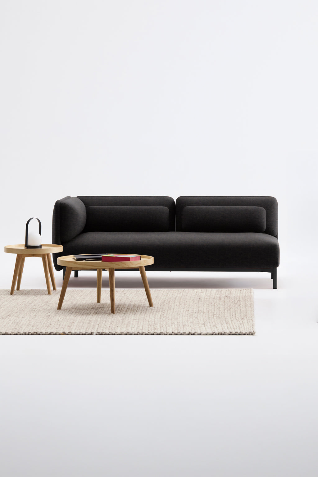 Un salon moderne présente le Canapé d'accueil modulaire Aurea - 1 accoudoir de Monsieur Bureau, un canapé noir disposé avec un tapis texturé beige clair, une table basse ronde en bois avec des magazines, et une table d'appoint à poignée sur un fond blanc.#finitions_noir