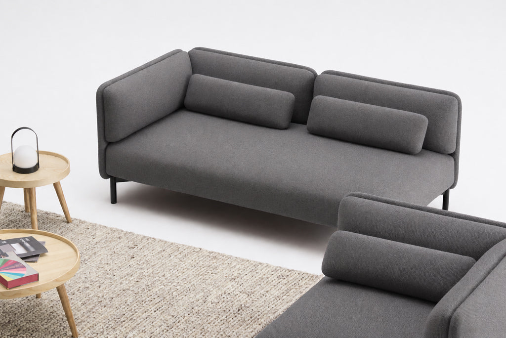 Un salon moderne et minimaliste présente le Canapé d'accueil modulaire Aurea - 1 accoudoir de Monsieur Bureau, associé à des coussins cylindriques, un tapis tissé léger et des tables gigognes rondes en bois surmontées de magazines et d'une lanterne décorative.