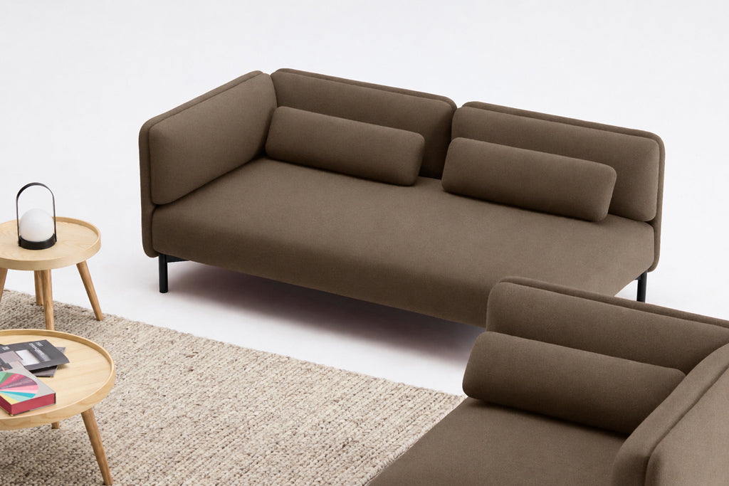 Le Canapé d'accueil modulaire Aurea - 1 accoudoir de Monsieur Bureau, conçu par Sascha Sartory, présente un look minimaliste brun moderne avec des coussins cylindriques, posés sur un tapis beige près d'une table d'appoint ronde en bois avec des livres et une lanterne.