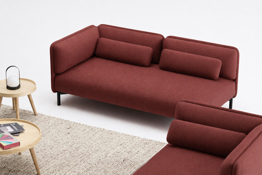 Un salon moderne avec le Canapé d'accueil modulaire Aurea - 1 accoudoir de Monsieur Bureau, un canapé rouge avec des coussins cylindriques, un tapis beige texturé, une table d'appoint ronde en bois avec une lanterne à bougie, et une pile de livres et de magazines.