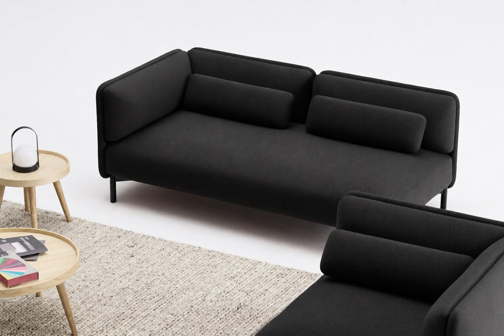 Un salon moderne avec un Canapé d'accueil modulaire Aurea gris foncé - 1 accoudoir de Monsieur Bureau, des coussins cylindriques, une table d'appoint en bois clair avec une lampe et des magazines, et un tapis beige texturé sur un sol blanc.