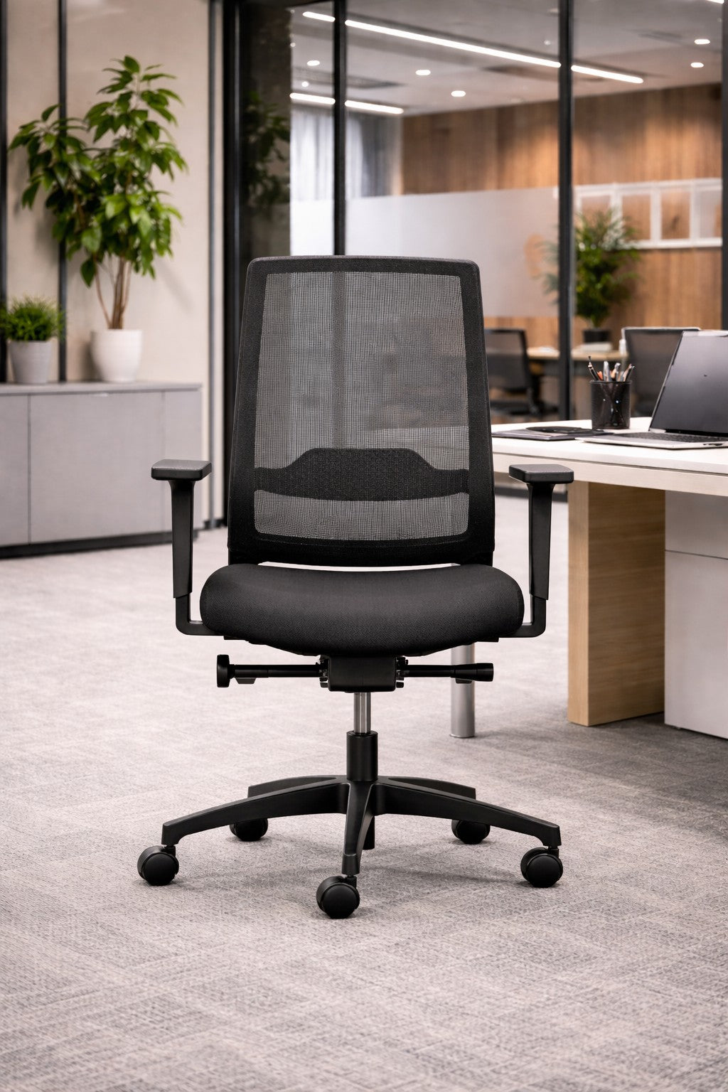 Un Fauteuil ergonomique professionnel Uneo de Monsieur Bureau, doté d'accoudoirs réglables et d'un revêtement en résille noire, est posé sur une moquette grise dans un bureau moderne, près d'un bureau avec un ordinateur portable, des articles de papeterie et des plantes en pot à l'arrière-plan.