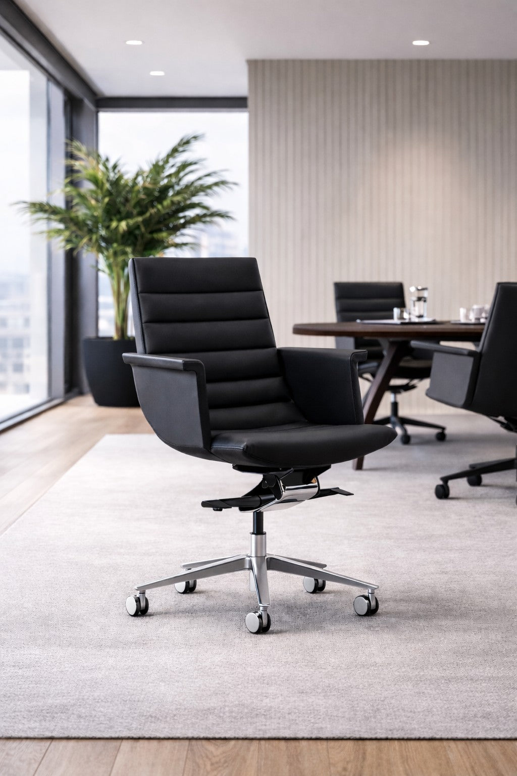 Le fauteuil design Altara dossier moyen & accoudoirs confort de Monsieur Bureau, noir avec base chromée et roulettes, est posé sur un tapis gris dans un bureau lumineux au sol en bois, avec grandes fenêtres, plante et table de conférence.