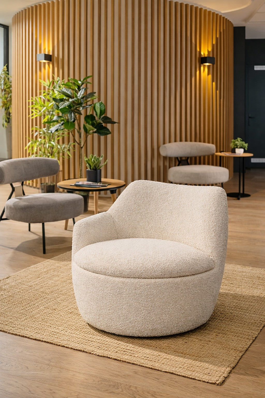 Un salon moderne présente la Chauffeuse d'accueil Kurl tissu bouclette de Monsieur Bureau en crème sur un tapis tissé, avec des chaises grises, des petites tables avec des plantes et un mur d'accent en lattes de bois avec des lumières pour une ambiance confortable et élégante.