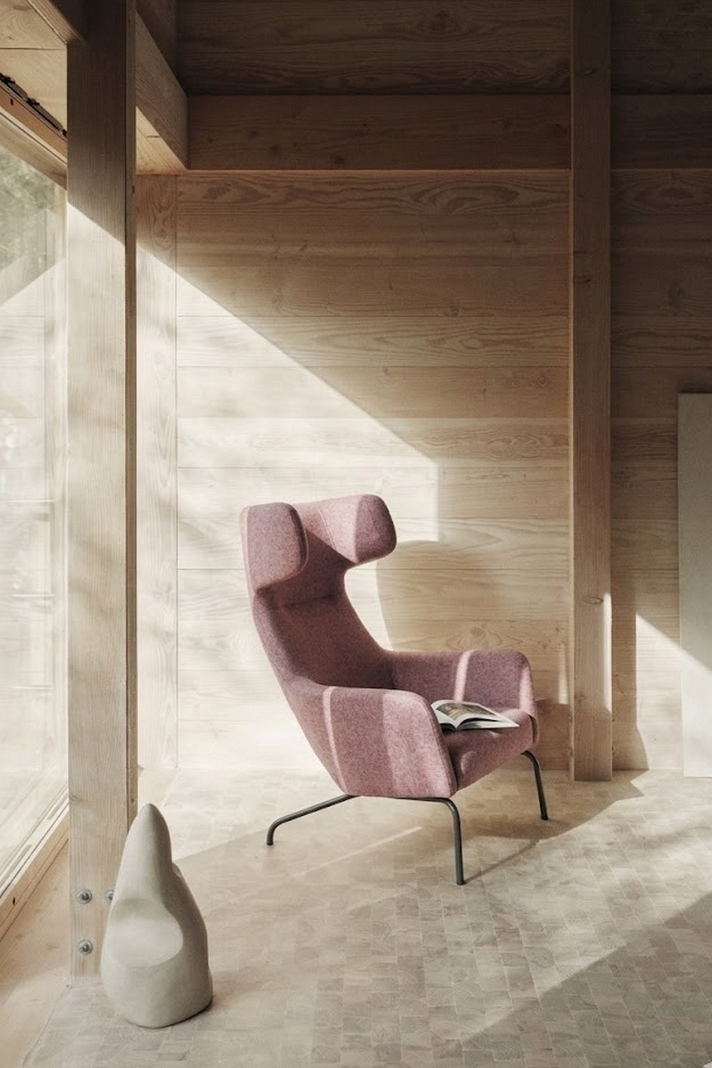 Le dossier Fauteuil lounge Fjord haut de Monsieur Bureau se trouve dans une pièce en bois éclairée par le soleil et au décor minimaliste. Une sculpture abstraite en forme de poire se trouve à proximité, tandis qu'un magazine ouvert est posé sur la chaise et que la lumière pénètre par une grande fenêtre.#finitions_vieux_rose