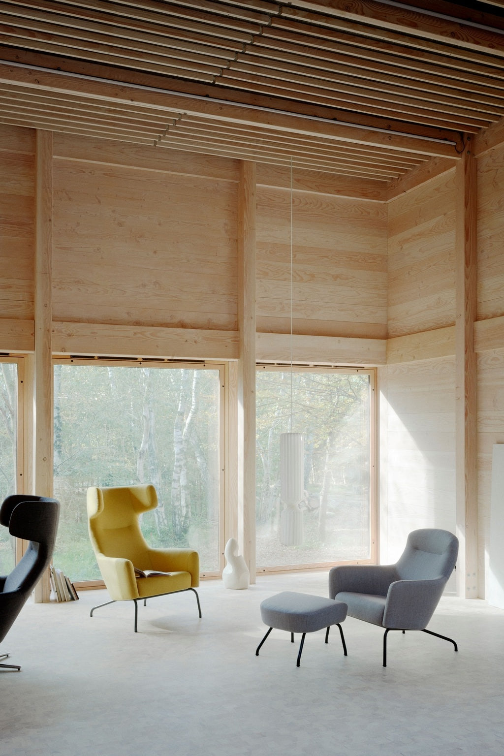 Une pièce moderne et minimaliste avec de grandes fenêtres, des panneaux de bois clair, un dossier jaune Fauteuil lounge Fjord haut de Monsieur Bureau, une chaise longue et un ottoman gris, un décor sculptural, une lampe suspendue et des arbres à l'extérieur pour les vibrations du design scandinave.
