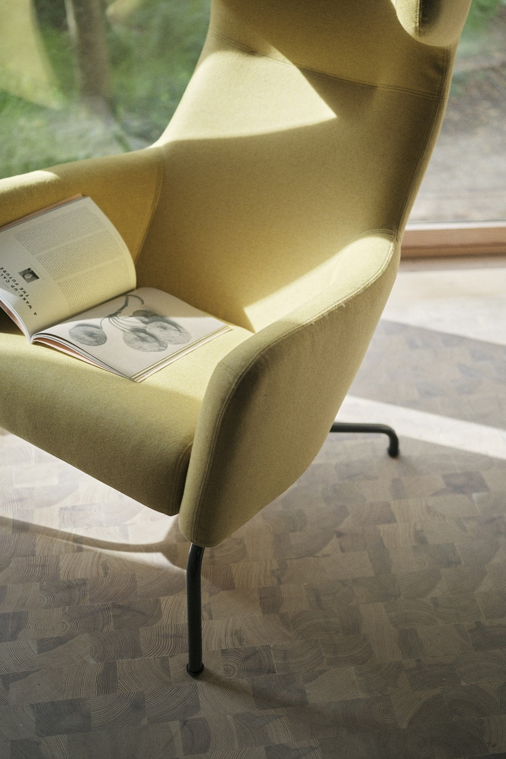 Le Fauteuil lounge Fjord haut dossier de Monsieur Bureau, en jaune moderne avec des pieds noirs, est assis près d'une fenêtre lumineuse. Un livre ouvert illustré repose sur son siège tandis que la lumière du soleil projette des ombres sur le sol en bois à motifs, évoquant le design scandinave.