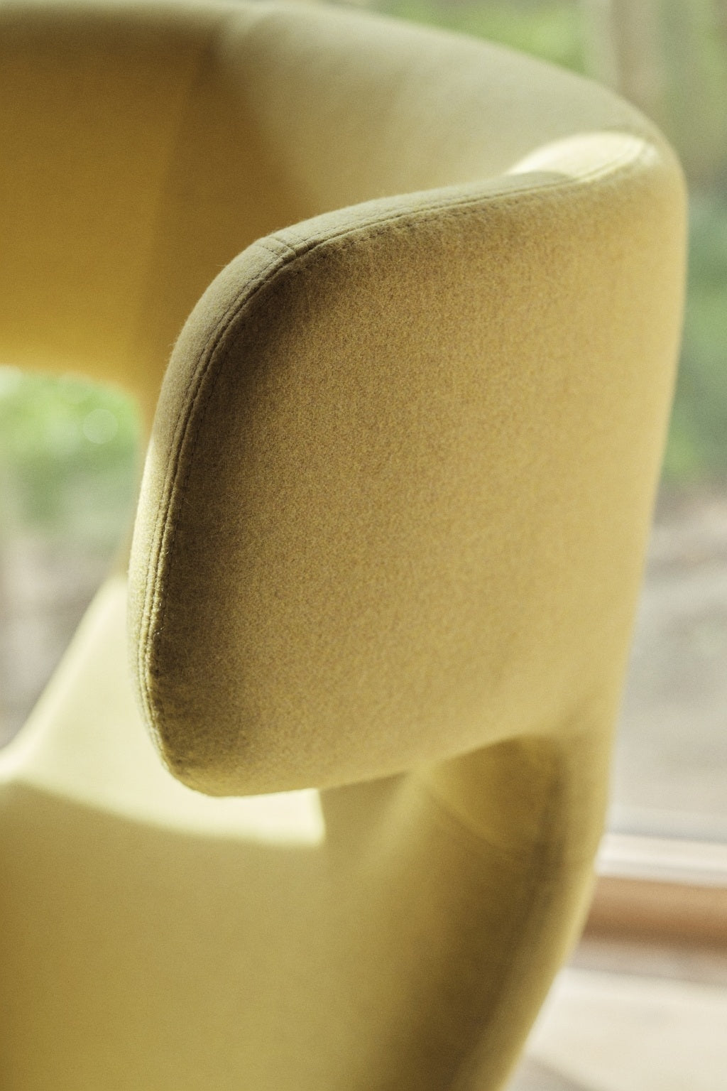 Gros plan sur l'accoudoir et le dossier incurvés du Fauteuil lounge Fjord haut dossier de Busk+Hertzog pour Monsieur Bureau, en tissu jaune. La lumière douce du soleil met en valeur sa texture, avec une fenêtre et de la verdure en arrière-plan pour un effet scandinave.