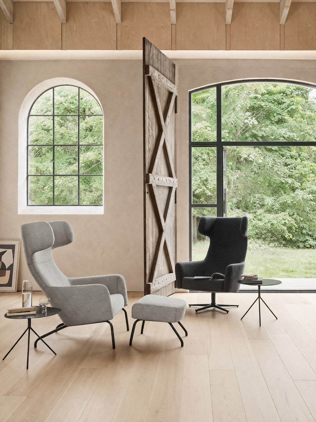 Salon moderne avec de grandes fenêtres arquées et rectangulaires, comprenant le Fauteuil lounge Fjord haut dossier de Monsieur Bureau (design Busk+Hertzog) en gris, un fauteuil noir, un ottoman, des tables d'appoint avec des verres, et une cloison en bois - le design scandinave brille.