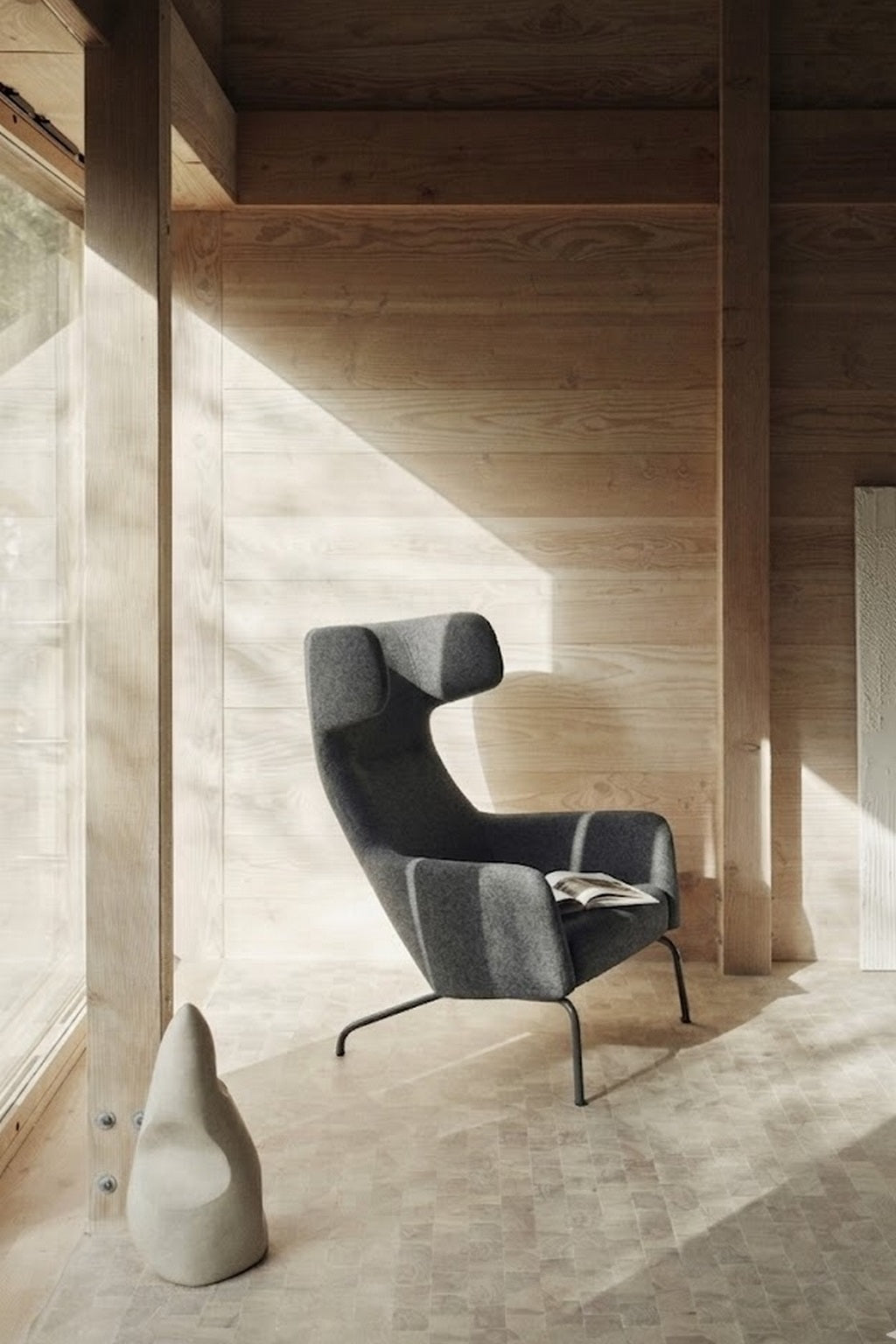 Le Fauteuil lounge Fjord haut dossier de Busk+Hertzog pour Monsieur Bureau, d'un gris moderne, est exposé à la lumière du soleil sur un sol en bois clair ; un magazine est posé sur son siège et une sculpture abstraite à proximité met en valeur la simplicité scandinave.#finitions_anthracite