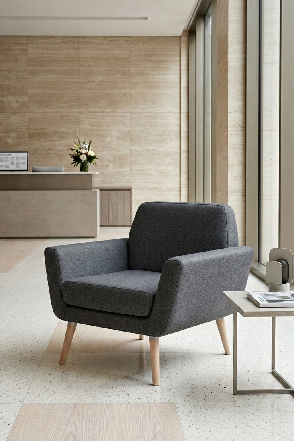 Le Fauteuil scandinave Natt de Robert ZOLLER de Monsieur Bureau, avec ses pieds en bois, se trouve à côté d'une table d'appoint dans un hall d'entrée minimaliste aux murs beiges, avec de grandes fenêtres et un bureau d'accueil avec des fleurs en arrière-plan.#finitions_anthracite