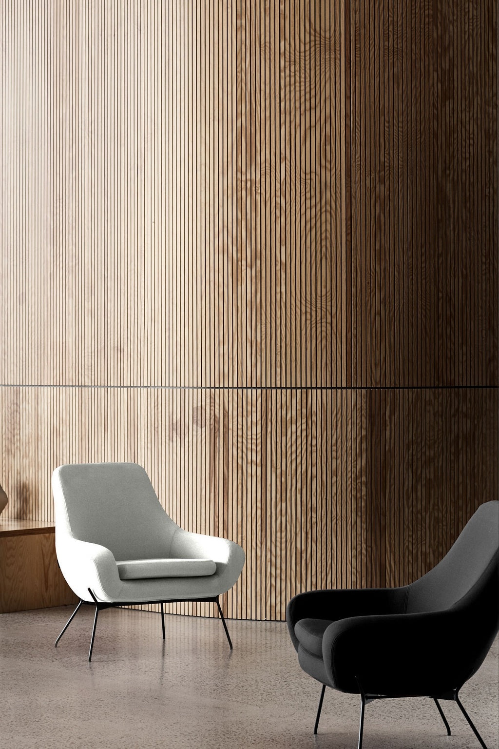 Une pièce moderne et minimaliste présentant le Fauteuil Lounge Nomi gris clair de Susanne GRØNLUND (Monsieur Bureau) et un fauteuil noir sur un sol poli, encadré par un haut mur de lattes en bois - idéal pour le design contemporain et le mobilier professionnel.